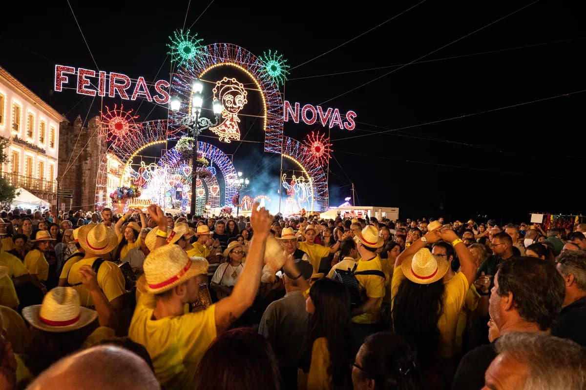 夜裡一座巨大的燈飾拱門寫著 FEIRAS NOVAS，中央有一張發光的人臉圖案，兩側是星形燈飾，底下擠滿戴草帽穿黃衣的人群，有人高舉雙手