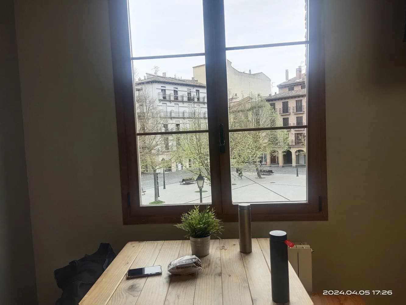 旅館窗邊的木桌，桌上兩支水壺和一盆小植物，窗外是城市廣場